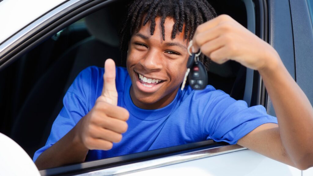 Happy young man in car holding keys and giving thumbs up.